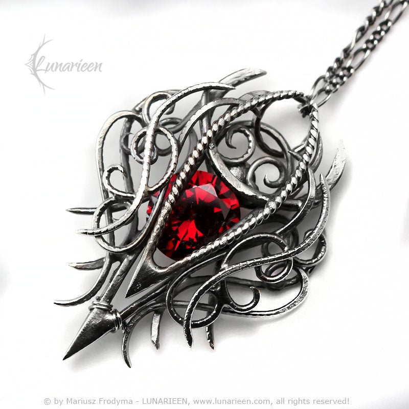 May include: A silver pendant necklace with an intricate, openwork design. The pendant features a large, faceted, red gemstone at its centre. The necklace has a delicate chain and a pointed, spear-like element at the bottom.